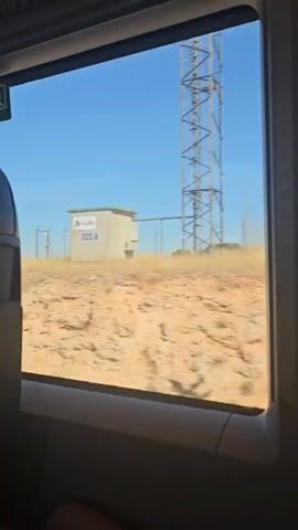 The video shows a view from a train window. The landscape outside is dry and arid, with sparse vegetation and a clear blue sky. The train appears to be moving, as indicated by the blurriness of the scenery. There are no people visible in the frame, except for a leg at the bottom of the screen. The interior of the train is dimly lit and out of focus. The video is short, lasting only a few seconds, and captures a fleeting glimpse of the passing landscape.