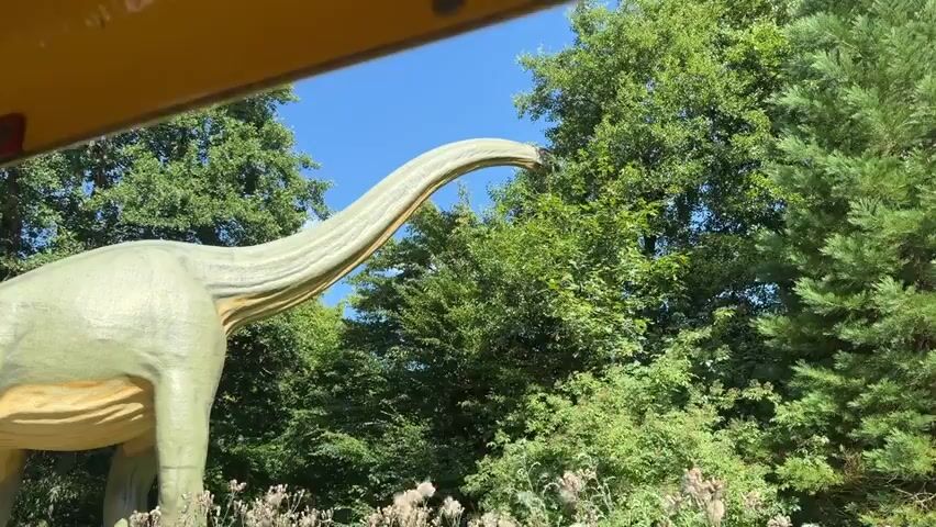 The video shows a close-up shot of a light green dinosaur statue. The statue is in a park or outdoor setting with lush green trees and a blue sky in the background. The camera appears to be moving, possibly from inside a vehicle or a moving attraction, as evidenced by the blurred yellow and red frame at the top of the image. The dinosaur statue has a long neck and tail, and its body is textured to resemble skin. The lighting suggests it is daytime.