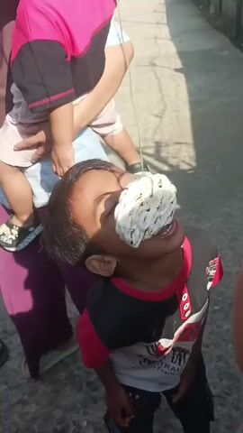 A young boy is attempting to eat a large, white cracker that is suspended from a string attached to his forehead. The cracker is positioned directly in front of his face, and he is tilting his head back and opening his mouth to try and bite it. He is wearing a black and red shirt with printed graphics. He is standing on a concrete surface that appears to be a walkway or street. There are other people around him, including someone in a purple outfit holding a small child. The environment looks like a residential area. The lighting indicates it is daytime.