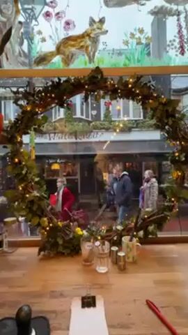 The video shows a view from inside a building, looking out through a window decorated with a large, circular wreath made of branches, leaves, and small lights. Above the wreath, the window is painted with nature scenes including a fox and an owl. Outside, the street is visible, with people walking by, some holding umbrellas in what appears to be rainy weather.  Across the street, a building with the sign "The White Rabbits" is visible. In the foreground, on a wooden surface, there are glasses, salt and pepper shakers, a mug, a clipboard with papers, and a red pen. The camera remains still throughout the video.