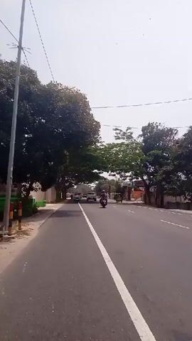 A short video shows a view from a vehicle moving down a road lined with trees and buildings. Several cars and motorcycles are visible on the road ahead. The sky is bright, suggesting daytime. The video appears to have been recorded from a handheld device.