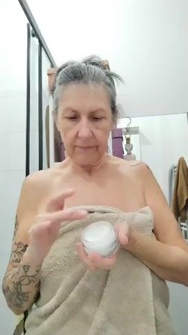 A woman in a bathroom is applying facial cream. She is fair-skinned with gray hair pulled up in a bun and a decorative tattoo on her left arm. The woman is standing, wrapped in a light brown towel covering her chest. She uses her right hand to take facial cream from a small white container and her left hand to apply it to her face and neck. A shower enclosure and a mirror with a towel bar and towel are behind her. The room appears to be a typical bathroom with white tiles. The product container appears to be L’Oreal Paris COLLAGEN.