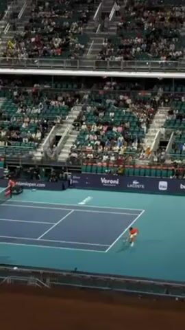 The video shows a tennis court in an indoor arena, likely during a match. The court is blue with white lines. A man in an orange shirt is standing on the court, presumably a player. The stands are mostly full with people watching the game. There are advertisements along the side of the court for brands like Lacoste and ITAU. The overall environment is that of a professional tennis event.