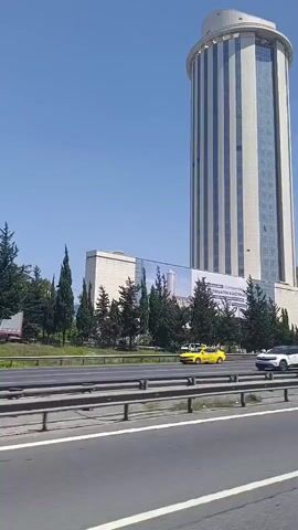 A tall, cylindrical building with a light-colored facade and vertical blue glass panels dominates the frame. A banner with Turkish text is visible on the lower section of the building. In front of the building, there is a row of green trees and a multi-lane road with a metal guardrail. Several vehicles, including cars, a white van, and a yellow and black bus, are moving on the road. The sky is clear and blue. The video appears to be filmed from a moving vehicle.
