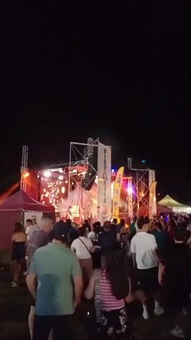 The video shows a crowd of people at an outdoor nighttime event. There is a stage with lights and speakers. People are standing and walking around. The ground appears to be grass.