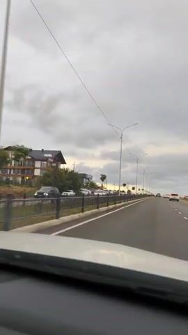 The video shows a view from inside a vehicle looking out towards a highway. On the left side of the highway, there is a multi-story hotel with a sign that says "HOTEL". In front of the hotel and along the side of the road, there is a row of parked cars. The highway itself has multiple lanes and is lined with streetlights. The sky is cloudy, suggesting it may be overcast or late in the day. The camera is positioned to capture the road ahead and the scenery to the left.