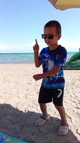 A young boy with sunglasses and a blue t-shirt with a picture of Cristiano Ronaldo and the Real Madrid crest is standing on a sandy beach. He is wearing black shorts and grey crocs. He is holding his right index finger up in the air and appears to be dancing or posing. In the background, there is the ocean, a beach umbrella, and other people in the water and on the shore. The sky is blue with some clouds. The video is shot in a landscape orientation.