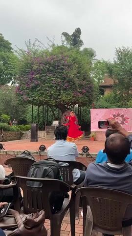 The video shows a woman performing a dance on a stage in front of an audience. She is wearing a red dress and gold jewelry, and her hair is styled in a bun. The stage is made of brick, and there is a large tree with purple flowers behind her. The audience is seated in brown plastic chairs. Many people are taking pictures and videos. There is a banner on the right side of the stage. The overall setting appears to be an outdoor cultural event.