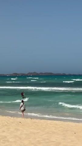 This media file captures a sunny day at the beach in La Oliva, Spain. The scene is dominated by a wide expanse of sandy beach in the foreground, leading to vibrant turquoise waters that are active with waves. In the middle ground, a kitesurfer is actively engaged in their sport. They are riding on a board and being propelled by a large, red and white kite that is visible in the sky. The kitesurfer appears to be wearing a dark wetsuit and a life vest. Further out in the ocean, a person can be seen walking near the water's edge on the beach. In the background, rolling hills or mountains form the coastline, suggesting a somewhat arid landscape. The sky is a clear, bright blue, indicating good weather conditions. The time of day appears to be daytime, likely mid-morning or afternoon, given the brightness of the sunlight. The overall mood is one of recreation and enjoyment of a beautiful coastal environment. The red and white kite has a visible logo with some text and the number "10" on it, but it is not clearly legible.