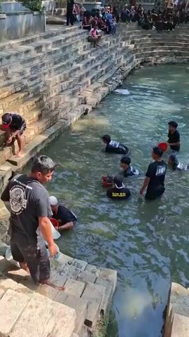 A high-angle shot captures an outdoor scene with a pool of murky water, surrounded by tiered stone steps. Several young people, predominantly male, are in the water, some holding red buckets. One person on the steps holds a white cloth. On the upper levels of the steps, a crowd of onlookers, many dressed in black, are seated. The environment suggests a natural spring or a traditional gathering place. The overall atmosphere appears to be one of activity and communal engagement.