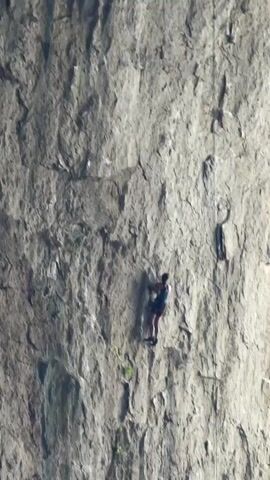 A person rock climbs up a sheer rock face. The climber, wearing a dark top and red shorts, is positioned in the center of the frame and is climbing upward on the textured, gray rock. A thin rope is visible, extending from the climber upwards and to the right. The background is dominated by the vast, uneven surface of the rock face, with various crevices and ledges. Some patches of green vegetation are sparsely scattered on the rock. The lighting suggests it is daytime, with the sun illuminating the scene from the side, creating subtle shadows.