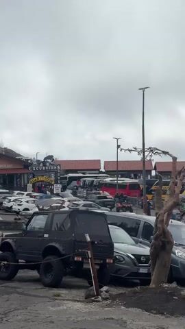 The video depicts a parking lot filled with various vehicles under an overcast sky. A black SUV with large tires is parked prominently in the foreground, with a dark gray Nissan SUV alongside it. Behind these vehicles, a building houses establishments like a "Snack Bar," "Tavola Calda," and "Sagonal," as well as "QUAD TOURS" excursions. People are milling around in front of the building. Further back, buses and more cars are visible, and a path leads upwards towards a dark, volcanic slope. The overall atmosphere is bustling, with signs of tourism and recreational activities evident in the parking area.
