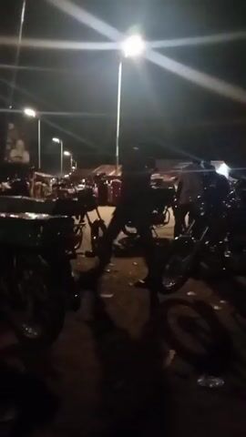 The video shows a night scene, likely outdoors, with multiple motorcycles parked. Several people are visible, though their faces are not clear due to the low light and motion blur. Some individuals appear to be standing near the motorcycles, while others are seated. In the background, there are structures that suggest stalls or shops, with lights illuminating the area. The ground is uneven and appears to be dirt or gravel, with some litter scattered around. The overall atmosphere is busy, possibly indicating a market or gathering. The camera is in motion throughout the clip, causing significant blur and making it difficult to discern specific details.