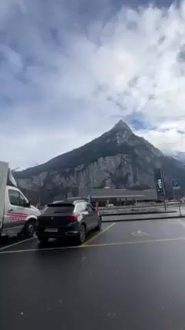 The video shows a parking lot in front of a gas station with a large mountain in the background. The parking lot is mostly empty, with a few cars parked in designated spots. The ground is wet, suggesting it may have recently rained or snowed. The gas station has a modern design with a flat roof and several gas pumps. In the background, a towering mountain dominates the landscape. The mountain is mostly covered in grey rock, with some patches of snow near the top. The sky is filled with clouds, giving the scene a somewhat overcast appearance.