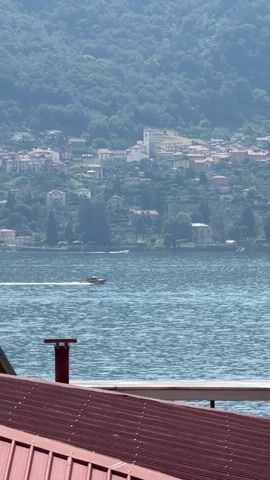 A boat travels across a body of water, leaving a wake behind it. In the background, a village with red-roofed houses and a white church is nestled on a hillside covered in trees. In the foreground, a portion of a red tiled roof and a red chimney are visible. The sky is hazy and appears to be daylight.