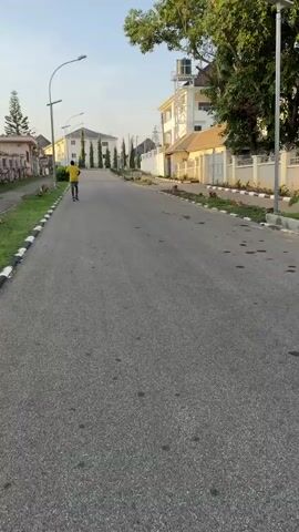 A man in a yellow shirt and gray shorts is running down an asphalt road in a residential area. He is moving away from the camera, on the right side of the road. The road is lined with grass and curbs painted in black and white. Houses are visible on both sides of the road. There are tall trees and bushes. Street lamps are spaced along the side of the road. The sky is clear and bright.