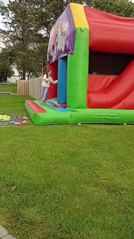 Two young girls are playing on an inflatable bouncy castle set up on a grassy lawn in front of a wooden fence. The bouncy castle is primarily red, green, and purple, with a large red slide. The girls appear to be interacting with each other, one is standing on the edge of the bouncy castle, while the other is inside it, reaching out to her. They are both wearing casual clothing. In the background, there are trees and a cloudy sky, suggesting an overcast day. Several pairs of shoes and a yellow mat are scattered on the grass near the bouncy castle. The overall scene depicts a playful outdoor activity, likely a birthday party or a similar event.