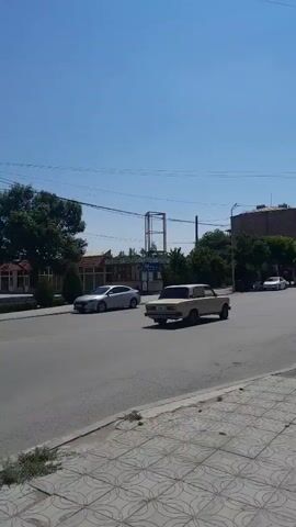 The video begins with a dark sedan driving past a light pole in an urban setting with buildings and trees in the background. The camera then pans to the left, showing a traffic light and a body of water. Another pan to the left reveals a white SUV crossing a pedestrian crossing in front of a building adorned with a large mural. The camera continues to pan, showing a white van and several other cars on a street with buildings on either side. The video concludes with a view of the street from a pedestrian's perspective, with cars driving by and buildings lining the road. The overall scene is an ordinary urban environment on a clear, sunny day.