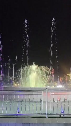 The video shows a nighttime view of a fountain with colorful lights. The water jets shoot up into the dark sky, illuminated by changing colored lights that shift between white, pink, and purple hues. The fountain is surrounded by a pool of water with a metal railing in the foreground. In the background, there are lights that suggest buildings or streetlights, but they are out of focus and indistinct. There are no people visible in the video. The overall impression is one of a public space at night, likely a park or plaza, featuring a decorative water display.
