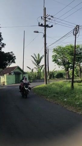 The video shows a rural scene. In the foreground, a paved road curves to the right. On the road, a motorcycle is moving towards the camera. The motorcycle carries two people, one in the front driving and one in the back. Both are wearing head coverings. To the left of the road, there is a small, green building with a brown roof, a small window, and a door. Behind the building, there are trees and a banana plant. In the middle ground, a utility pole with many wires is prominent. There is also a smaller pole behind it. The background consists of green fields and trees under a clear blue sky. The lighting suggests it is daytime, likely morning or late afternoon due to the quality of light.