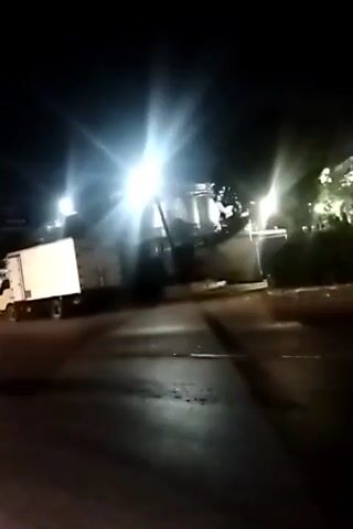 The video shows a night-time scene with multiple bright lights illuminating the area. A white truck is parked on the left side of the road. The road appears to be paved, and there is some debris scattered on it. The environment seems to be an urban or industrial area, with buildings and trees visible in the background. The camera is moving, suggesting it might be recording from a vehicle. The overall impression is a dimly lit, somewhat blurry scene at night.