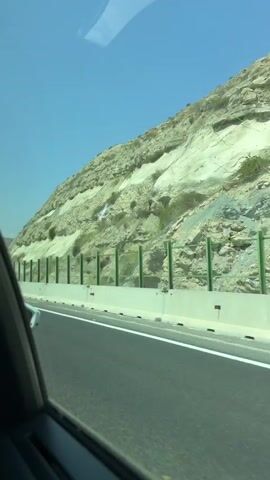 The video shows a view from inside a car traveling on a highway next to a rocky hillside. The hillside is layered with light-colored rock and some sparse vegetation. A green metal fence runs along the edge of the road, separating it from the hillside. The road is paved and has a white line marking the lane. The sky is clear and blue. The camera is positioned to the right side of the vehicle, and part of the car's interior, including a window frame, is visible in the foreground. The car appears to be moving at a moderate speed.