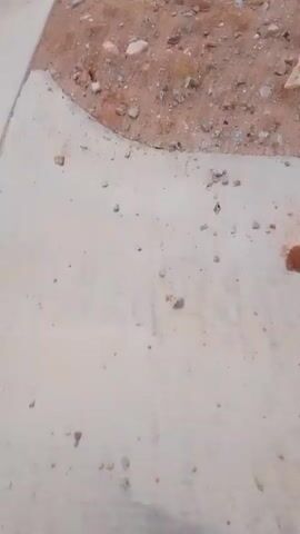 The video begins with a close-up of a concrete curb and a patch of disturbed earth with scattered rocks and debris. The camera then pans to the right, revealing a street under construction with more dirt, rocks, and some discarded wooden planks. In the background, there are stacks of building materials, possibly tiles or pavers. As the camera continues to move, the scene broadens to show a partially built house or development, with a modern architectural style featuring a pitched roof, large windows, and a gated entrance with a guardhouse. The area surrounding the house is still a construction site, with unpaved ground and unfinished landscaping. The sky is overcast, suggesting it may be a cloudy day.