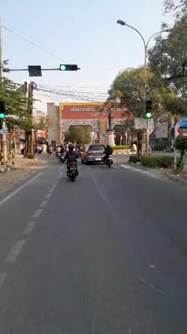 The video captures a street scene in front of a large archway bearing the text PEMERINTAH KABUPATEN TEGAL. Several vehicles, including motorcycles and a silver minivan, are present on the road. Traffic lights are visible, and some are illuminated green. People are seen riding the motorcycles. The archway is adorned with decorative patterns and flags. A statue stands in front of the archway. The sky appears clear, suggesting daytime.