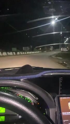 The video is filmed from the perspective of a driver inside a vehicle at night. The dashboard is visible in the foreground, with a steering wheel, instrument cluster, and a tablet mounted on the right. The instrument cluster displays green illuminated gauges and warning lights. The road ahead is illuminated by streetlights, which are visible in the distance. The surroundings are dark, with some buildings and signs visible along the roadside. The vehicle appears to be moving on a paved road, possibly in a rural or suburban area. The overall atmosphere suggests a night drive.