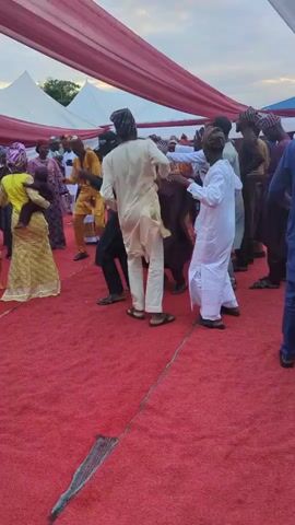 A low-angle full shot shows an outdoor gathering with people dancing on a red carpet under a white tent with a red and white ceiling. The people are dressed in traditional Nigerian attire. In the foreground, a man in a light yellow tunic and pants is dancing with a man in dark pants and a dark long-sleeved shirt. Another man in a white tunic and pants stands to the right of them. In the background, other people are dancing. To the left, a woman in a yellow dress holds a baby. The ground under the red carpet is visible in the foreground.