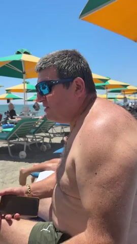 A middle-aged, gray-haired man is sitting on a beach lounge chair with his shirt off, wearing sunglasses and a gold chain around his neck. He’s holding a dark smartphone in his left hand and wearing a watch on his left wrist. The scene takes place on a beach with multiple rows of lounge chairs and colorful beach umbrellas in the background. The man looks towards the sea and appears to be speaking. The sky is clear and bright. There are other people sunbathing on the lounge chairs in the background.