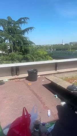 The video shows an outdoor setting on a flat rooftop with a view of the Eiffel Tower in the distance. The roof is littered with trash, including plastic bottles, bags, and discarded food containers. A black trash can is situated near the edge of the roof. A man wearing a pink t-shirt and gray shorts walks over to a charcoal grill and attends to it. The foreground includes a red shopping bag and a half-filled glass. A bridge with streetlights and passing vehicles is visible in the middle ground. The sky is clear and blue.