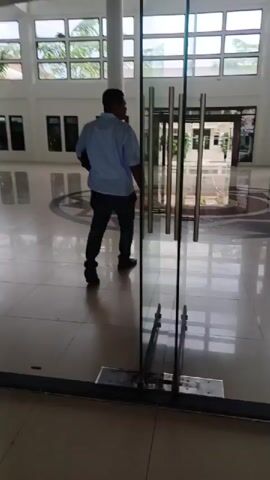 A man in a light blue, short-sleeved shirt and dark pants walks from right to left across a large, brightly lit lobby. The floor is tiled and reflective, and the lobby has high ceilings with large windows and white columns. The man is walking at a moderate pace and looking around. There's a staircase in the background and glass doors in the foreground.