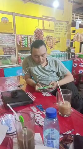 The video shows a man sitting at a red table with two glasses of brown liquid with straws and a bottle of blue-capped water. He is wearing a grey t-shirt with a dark pattern and has his hair pulled back in a bun. The man is looking down at his phone, which he is holding in his hands. To his left, there is a laptop with a black mouse. The table has various items on it, including what appears to be a menu or placemat with yellow and orange accents. In the background, there are shelves stocked with snacks and drinks, and the walls are painted yellow. There is a light fixture hanging from the ceiling. The overall impression is a casual setting, possibly a roadside eatery or a small shop.