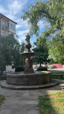 A stone fountain with a statue at the top sits in a paved area surrounded by greenery. To the left, a modern building with balconies is visible. To the right, trees and a glimpse of a red-tiled area with people and bins suggest a public space. The sky is blue with some clouds. A person walks past the fountain in the background.