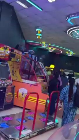 The video shows a brightly lit arcade with various game machines and people. There's a basketball game, a dancing game with a metal platform, and a clear plastic ball machine with colorful lights. People are walking around, and some are playing the games. The arcade has a modern design with neon lights and a patterned ceiling. The camera moves through the arcade, capturing the bustling atmosphere.