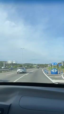 The view from the dashboard of a car traveling down a multi-lane highway. The sky is blue with scattered clouds. Cars are traveling in both directions. Streetlights line the highway. There are road signs to the right of the road, and a grassy median on the left. The car is playing music.