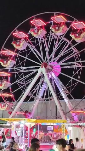 This is a vertical, low-angle shot of a Ferris wheel illuminated by red and pink lights at night. The Ferris wheel is large and white with many spokes. The gondolas, which are red and gold, are hanging from the edge of the wheel. Below the Ferris wheel, there are several carnival stalls. People are visible in the foreground and around the stalls. The background is dark, indicating it is nighttime. The Ferris wheel is slowly rotating, and some of the lights appear to be flashing.