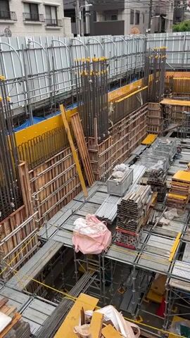 This is an aerial view of a construction site with an unfinished building. The area is filled with scaffolding, wooden forms, and rebar. In the center of the frame, there are stacks of lumber and metal pipes. A pink-wrapped bundle is also visible on a platform. Several large vertical rebar structures rise from the foundation. The environment appears to be an urban setting with buildings visible in the background.