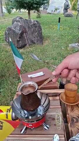 A close-up shot captures a hand holding a spoon over a pot of coffee brewing on a small camping stove. An Italian flag is planted in the ground next to the pot. In the background, a large rock and a statue are visible in a grassy area.