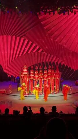 From a low angle, an audience watches a performance. The scene is illuminated by bright red lighting, and a large, multi-dimensional fan structure hangs above the stage. Several performers, dressed in colorful costumes depicting stylized animals, move around a formation of red and gold objects resembling miniature pagodas. In the background, other performers are visible, interacting with props. The audience members are visible in silhouette, some holding up phones to record the performance. The overall impression is a vibrant, stylized spectacle.
