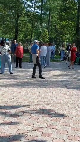 A group of people are gathered outdoors on a paved area in a park. The individuals appear to be engaged in some form of organized activity, possibly dancing or exercise, as several are in motion and facing a central point. The environment includes trees and a sunny sky, indicating daytime. Among the people, there are adults of varying ages, some dressed in casual attire and others in more sporty clothing. A pigeon is visible on the ground in the foreground. The overall scene depicts a public gathering in a park setting.