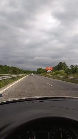 This is a first-person perspective video from inside a car driving on a two-lane highway under a cloudy sky. The road is paved and shows signs of wear. Guardrails line both sides of the road, with vegetation and trees growing beyond them. In the distance, a white car is visible in the same lane. A billboard with an advertisement is positioned on the right side of the road. The car's dashboard is partially visible in the foreground, including a speedometer that indicates a speed of approximately 100 km/h. The weather appears overcast and possibly stormy.