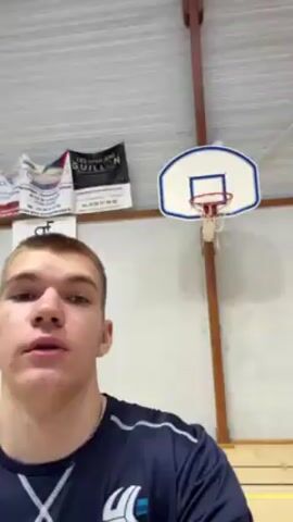 The video shows a young man in a blue athletic shirt, possibly filming himself in a gymnasium. Above him is a basketball hoop mounted on a white wall with promotional posters. He shoots a yellow ball at the hoop, and it goes in. Sounds of a bouncing basketball are audible.