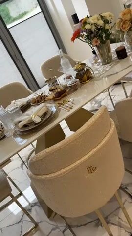 The camera pans across an elegantly set dining table, likely for a special occasion. The table is white marble with a gold geometric base, surrounded by plush, textured beige chairs with gold accents. The floor is a marble pattern. The table setting includes ornate gold-rimmed plates with white napkins held by gold rings. There are clear glasses and gold-colored cutlery. In the center of the table, there's a white runner with a floral pattern. Several dishes hold an assortment of cookies, candies, and pastries. A glass water bottle and a gold teapot are also present. Vases of colorful flowers add to the aesthetic. The scene suggests a high-end, luxurious dining setting. No people are visible.