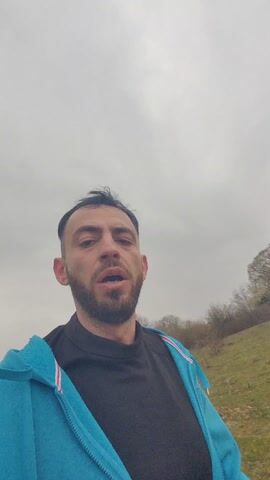 A man with dark hair and a beard is speaking to the camera while running. He is wearing a black shirt and a light blue zip-up hoodie. The background is a grassy field with trees and a cloudy sky.
