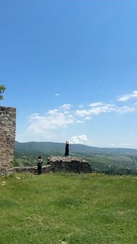 The video shows a landscape view with two people. In the foreground, there is green grass and a stone wall on the left. In the middle ground, there is a ruined stone structure where one person is standing. This person is wearing a black dress and white shoes, and they are holding their hands up to their face as if taking a photo. To the left of this person, on the same ruined structure, another person is standing and looking towards the landscape. This person is wearing a patterned shirt and jeans. The background consists of rolling green hills and a village nestled in the valley, all under a bright blue sky with scattered white clouds. The camera is positioned low, looking up slightly at the scene.