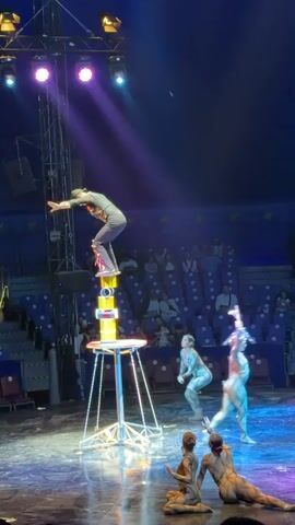 The video shows a circus performance in a large tent. A performer is balancing on top of a stack of cylinders on a table. Two other performers in light-colored costumes are on the stage floor, one standing and one kneeling. In the background, an audience is seated in bleachers. Spotlights illuminate the stage.