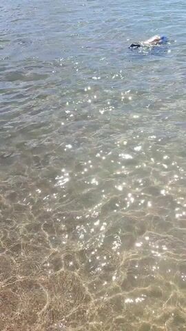 The video shows a crowded beach with many people swimming and playing in the water. The water is clear and shallow, with sunlight reflecting off the surface. In the foreground, a child wearing arm floaties sits in the water. To the right, a boy stands in the water, facing away from the camera. In the background, numerous people are visible in the water and on the shore. The sky is clear and blue.
