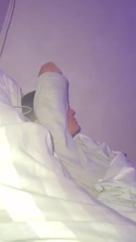 A close-up shot shows a young person in bed. They are lying on their side, partially covered by white striped bedding. The person is wearing a grey long-sleeved shirt. The room is dimly lit with a purple hue cast on the wall behind them. The person lifts their right arm, bending it at the elbow, and rests their hand on their forehead. They then lower their arm and turn their head slightly to look towards the camera. Their eyes are open and they have a neutral expression. The video is composed of a series of short, still frames edited together to create a short animation or GIF.